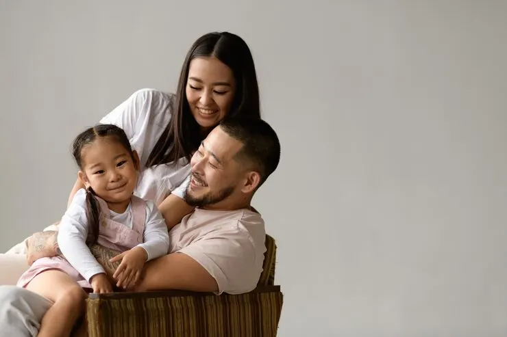 Happy asian parents looking toddler daughter while sitting armchair isolated grey